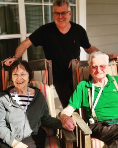 A senior husband and wife sit on the porch at Tamarisk Senior Living and their adult son stands behind them