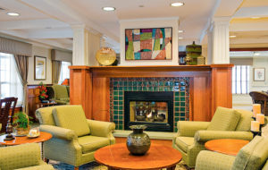 A cozy living room space with a large fireplace at the center of the image and four green armchairs arranged in front of the fireplace around a circular coffee table. A colorful piece of contemporary art hangs over the fireplace.