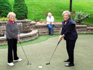 Three women together at the Tamarisk Senior Living putting green. Two are holding golf clubs while one sits watching, all are smiling at the camera.